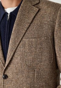 Blazer en tweed marron avec un revers cranté, une fermeture à un bouton et une poche poitrine. La texture est tissée avec un motif subtil.