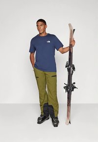 Άντρας με πράσινο σορτς σκι, μαύρες μπότες σκι και μπλε μπλουζάκι κρατάει ένα ζευγάρι μαύρες και καφέ σανίδες σκι, ενώ στέκεται μπροστά από απλό λευκό φόντο.