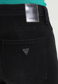 Jeans de denim negro con un parche plateado del logo de GUESS y una etiqueta triangular de GUESS en el bolsillo trasero. Textura suave con un ajuste ajustado.