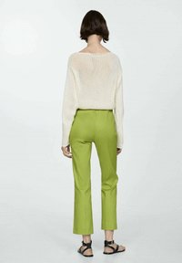 Off-white gebreide trui met wijde halslijn, gecombineerd met slim-fit, cropped groene broeken. Model draagt zwarte sandalen en kijkt van de camera weg.
