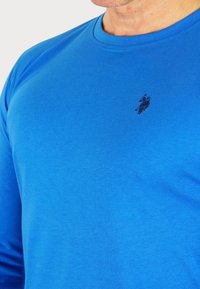Blauw langslavend shirt van zachte katoen, met een ronde halslijn en een klein zwart geborduurd logo op de borst.