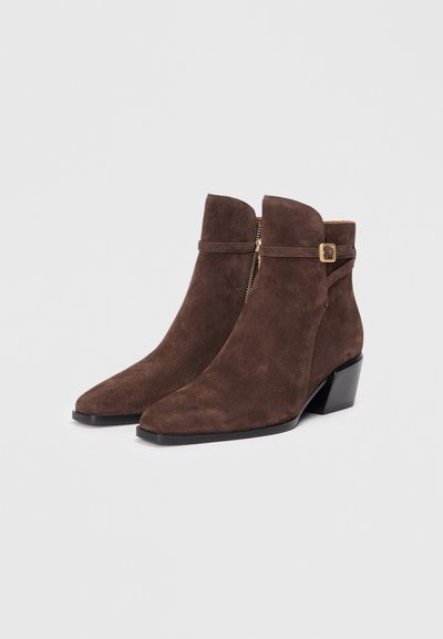 Bottines en daim marron avec un bout pointu, fermeture éclair sur le côté et un petit talon carré. Présente une sangle avec une boucle rectangulaire en accent.