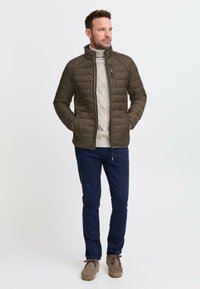 Mann i olivengrønn quiltet jakke over en beige kabelstrikket genser, marineblå bukser og brune semskede sko, stående med hendene i lommene.