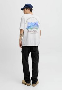 Balta grafiskā T-kreksa ar akvareļa kalnu dizainu aizmugurē un tekstu. Apģērbta kopā ar melniem kravas šortiem un bēšiem apaviem.