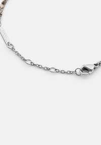 Ezüst lánc karkötő, kör alakú szemekkel és homárkapoccsal, „Daniel Wellington” felirattal ellátott címkével. Simább textúrával és fényes felülettel rendelkezik.