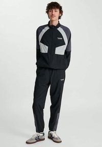 Jonge man die staat, gekleed in een zwart- en grijs trainingsjack met rits en bijpassende broek, met witte en grijze sneakers.