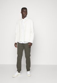 Chaqueta de mezclilla blanca con cierre de botones, llevada sobre una camisa clara, combinada con pantalones verde oliva y zapatillas blancas. Diseño simple y ajuste clásico.