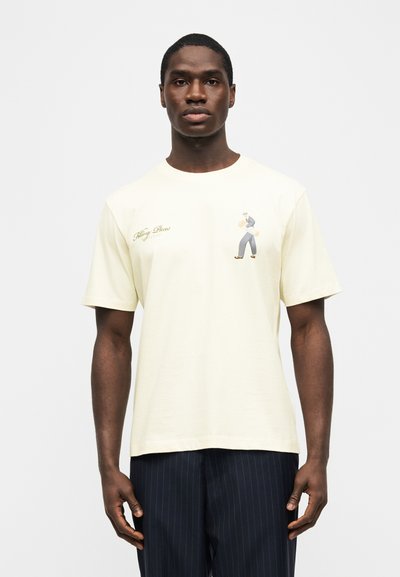 Άνδρας που φοράει εκρού κοντομάνικο μπλουζάκι με μικρό κείμενο και γραφικό από έναν πεζό στο στήθος, συνδυασμένο με σκούρο παντελόνι με ρίγες.