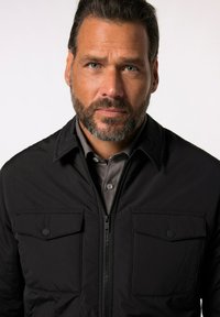 Schwarze Zip-Jacke mit Kragen, zwei Brusttaschen und glatter Textur. Über einem grauen Hemd mit Knöpfen getragen.
