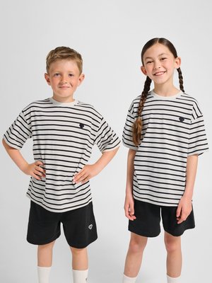 Dwoje uśmiechniętych dzieci stoi obok siebie, ubranych w pasujące białe i czarne koszulki w paski oraz czarne spodenki, jedno z rękami na biodrach, drugie z warkoczami.