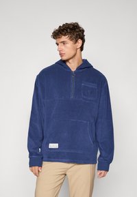 Felpa con cappuccio di colore blu navy in pile, con tasca frontale, colletto a mezza zip e logo ricamato. Presenta polsini a costine e un'etichetta sull'orlo.