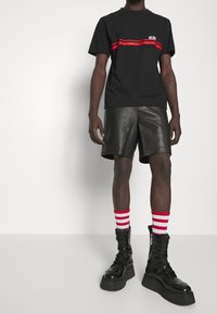 Camiseta de algodón negra con una franja roja, combinada con pantalones cortos de cuero negros y botas negras hasta la rodilla. Incluye calcetas a rayas rojas y blancas.