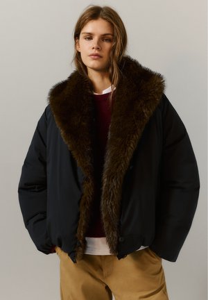 Jeune femme portant une veste noire avec un grand col en fourrure marron, un pull bordeaux, un sous-vêtement blanc et un pantalon beige, debout devant un fond uni.