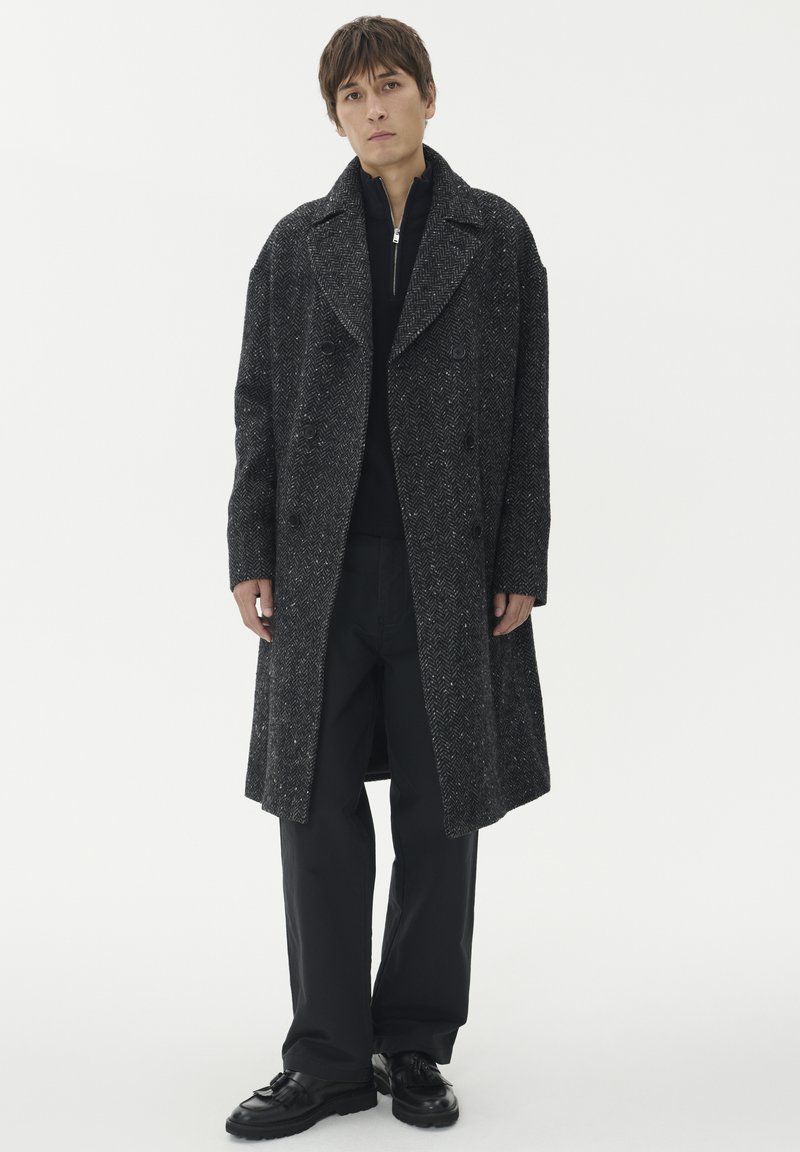 Giovane uomo in piedi che indossa un lungo cappotto a spina di pesce grigio scuro, un maglione nero con zip, pantaloni neri a gamba wide e scarpe in pelle nera.