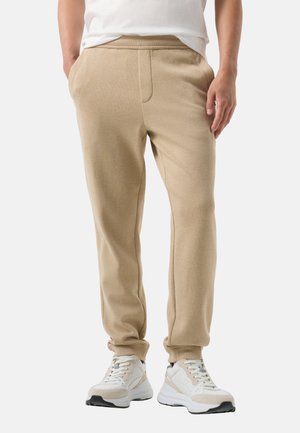Man draagt beige sweatpants en witte sneakers, staat met handen in de zakken tegen een effen achtergrond.