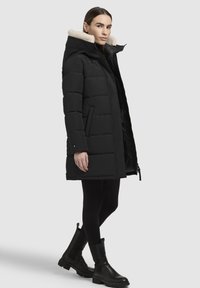 Schwarze Pufferjacke mit einem Kragen aus Kunstfell, gerader Schnitt, Reißverschluss und Seitentaschen. Ein Model trägt sie mit schwarzen Leggings und Stiefeln.