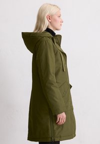 Olivgrön knälång kappa med huva, dragkedjeförsedda sidor och tryckknappsfickor. Slät textur och rak silhuett.