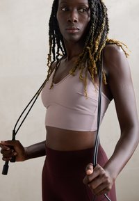 Reggiseno sportivo rosa chiaro con scollatura rotonda e spalline sottili, abbinato a leggings a vita alta di color marrone scuro, mentre tiene una corda per saltare.
