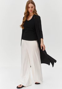 Czarny długi kardigan na czarnej bluzce i kremowych szerokich spodniach. Gładka tekstura; luźny krój. Sandały o minimalistycznym designie.