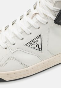 Sneaker bianco in pelle con dettagli neri, caratterizzato da un patch a forma triangolare, superficie texturizzata e lacci bianchi. Design classico alto.