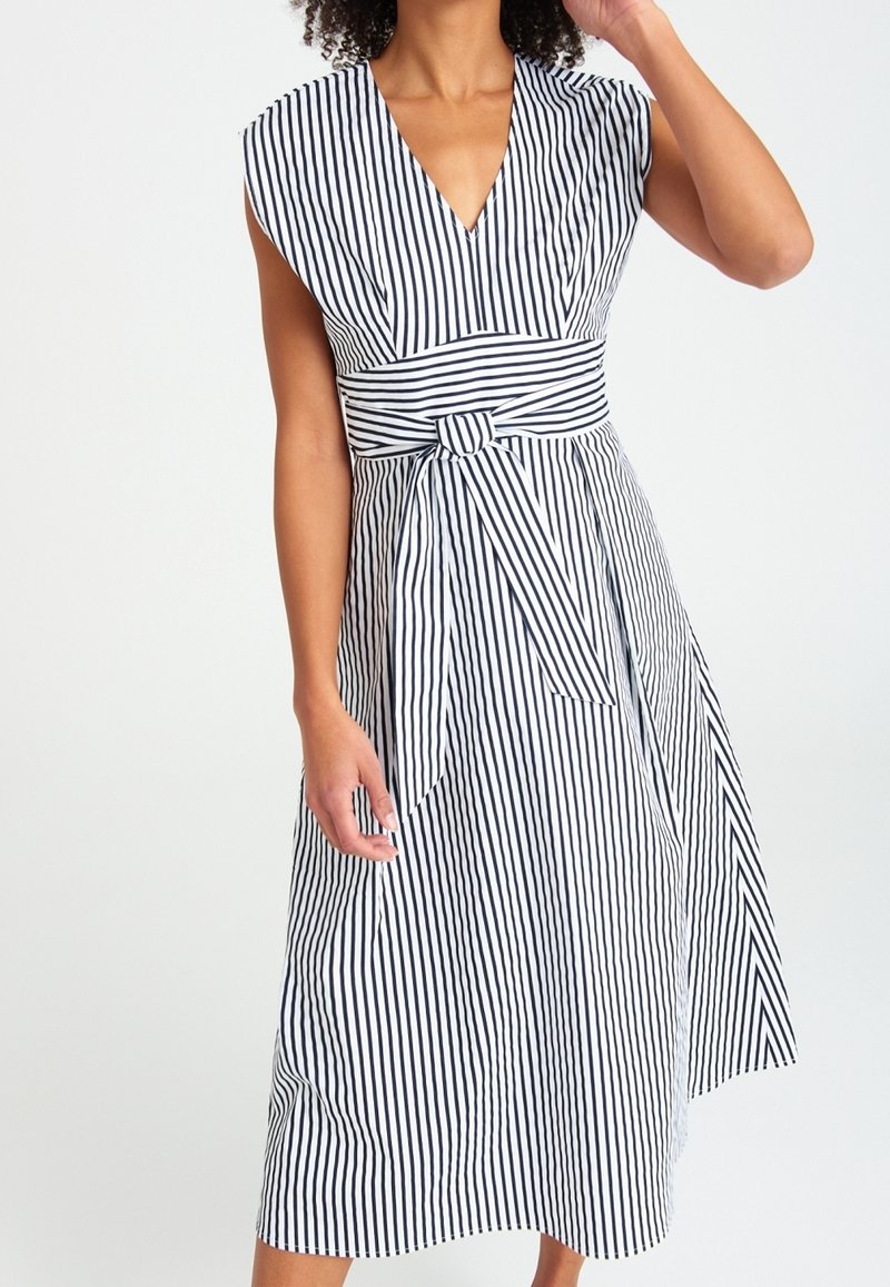 Femme portant une robe sans manches rayée noire et blanche à rayures verticales avec un décolleté en V profond et un nœud de ceinture rayé assorti.