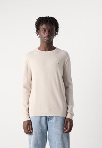 Beige textured knit sweater with long sleeves and round neckline. Subtle logo on the left chest. Paired with light blue denim jeans.