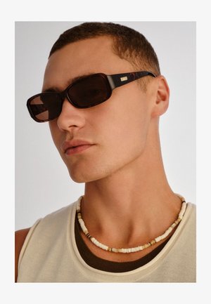 Noor mees kannab ruudukujulisi tumedaid päikeseprille, valget helmestega kaelakeed ning kihilist beeži ja pruuni varrukateta ülaosa.