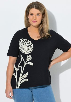 Frau mit langen blonden Haaren, trägt ein schwarzes kurzärmeliges Shirt mit großem weißem Blumenmuster und blaue Jeans, steht vor weißem Hintergrund.