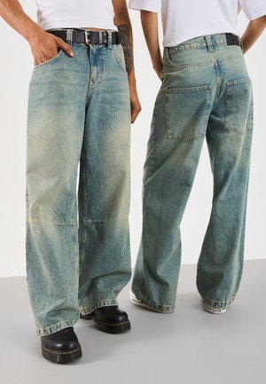 Dos personas que llevan vaqueros anchos azul claro y camisetas blancas, una mirando hacia adelante con botas negras y la otra de espaldas con zapatillas blancas.