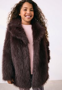 Manteau en fausse fourrure brun foncé avec un large col, manches longues et une texture pelucheuse ; porté par-dessus un haut rose et des leggings à motifs.