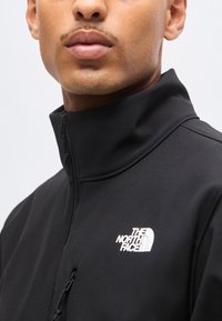 Gros plan d'une personne portant une veste noire zippée avec le logo The North Face sur la poitrine, sur un fond uni.