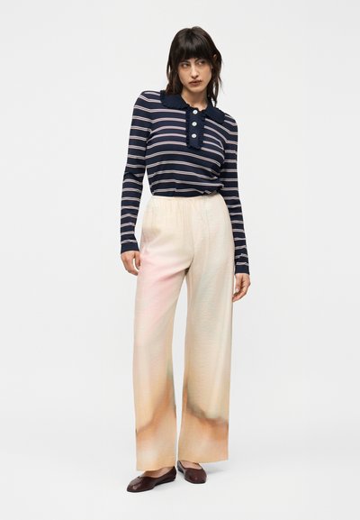 Γυναίκα στέκεται φορώντας μακρυμάνικη μπλούζα ναυτικού στυλ με ρίγες και κουμπιά, μαζί με εκρού παντελόνι φαρδύ με διακριτικό βαθμωτό σχέδιο, συνδυασμένο με σκούρα επίπεδα παπούτσια.