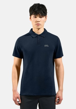 Jonge man met kort zwart haar, gekleed in een marineblauw poloshirt met een klein wit logo op de linkerborst, tegen een effen achtergrond.