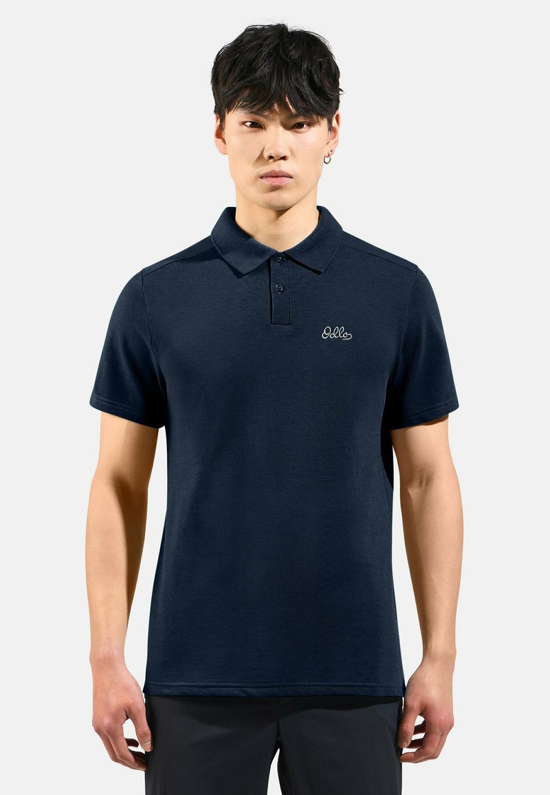 Junger Mann mit kurzem schwarzen Haar, der ein dunkelblaues Poloshirt mit einem kleinen weißen Logo auf der linken Brust vor einem einfarbigen Hintergrund trägt.