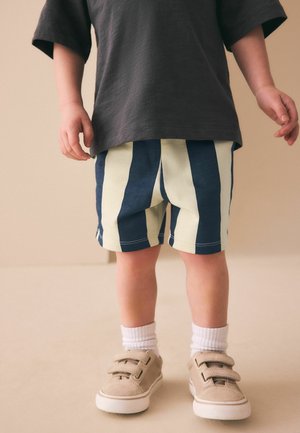 Småbarn iført marineblå og cremefarvede stribede shorts, mørk t-shirt, hvide sokker og beige sneakers står på en neutral gulv.