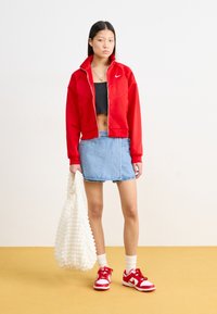 Punainen vetoketjullinen takki, musta crop-top, farkkuhame, valkoinen teksturoitu laukku ja punaiset sneakerit valkoisilla yksityiskohdilla. Malli seisoo keltaisella lattialla.