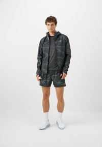Svart idrettsjakke med teksturert geometrisk mønster, kombinert med matchende shorts. Grå t-skjorte under, hvite joggesko og ankelstrømper.