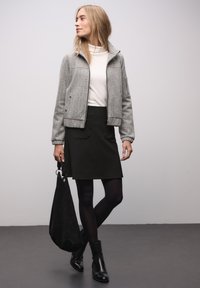 Šedá texturovaná bunda s žebrovanými manžetami, bílý rolák, černá sukně do áčka a černé kotníkové boty, držící černou semišovou tašku.