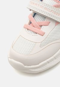 White athletic shoe with a textured mesh upper, pink accents, and a velcro strap. Features a rounded toe and rubber sole.