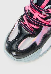 Nærbilde av en sneakers tå med iriserende svart overdel, neongrønne og svarte lisser, og lys blå såle.