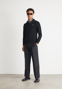 Mann trägt schwarze Sonnenbrille, einen schwarzen Pullover mit Viertel-Reißverschluss über einem gemusterten Hemd, schwarze Hose und schwarze Lederschuhe, stehend vor einer schlichten Wand.