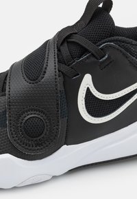 Chaussure de sport noire avec un dessus synthétique texturé, des accents en maille, un logo swoosh blanc, et une sangle Velcro sur les lacets pour un soutien.
