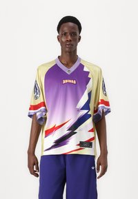 Lila és sárga póló, zig-zag mintával, rövid ujjakkal, V-nyakkal és Adidas logóval az elején. Kék rövidnadrággal párosítva.