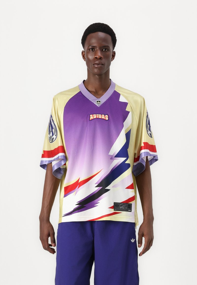 Lila és sárga póló, zig-zag mintával, rövid ujjakkal, V-nyakkal és Adidas logóval az elején. Kék rövidnadrággal párosítva.