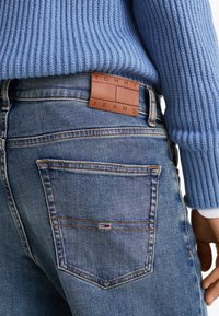 Jeans de denim con un lavado azul claro, que cuentan con un parche de cuero etiquetado "TOMMY JEANS" y bolsillos traseros clásicos con costuras naranjas.