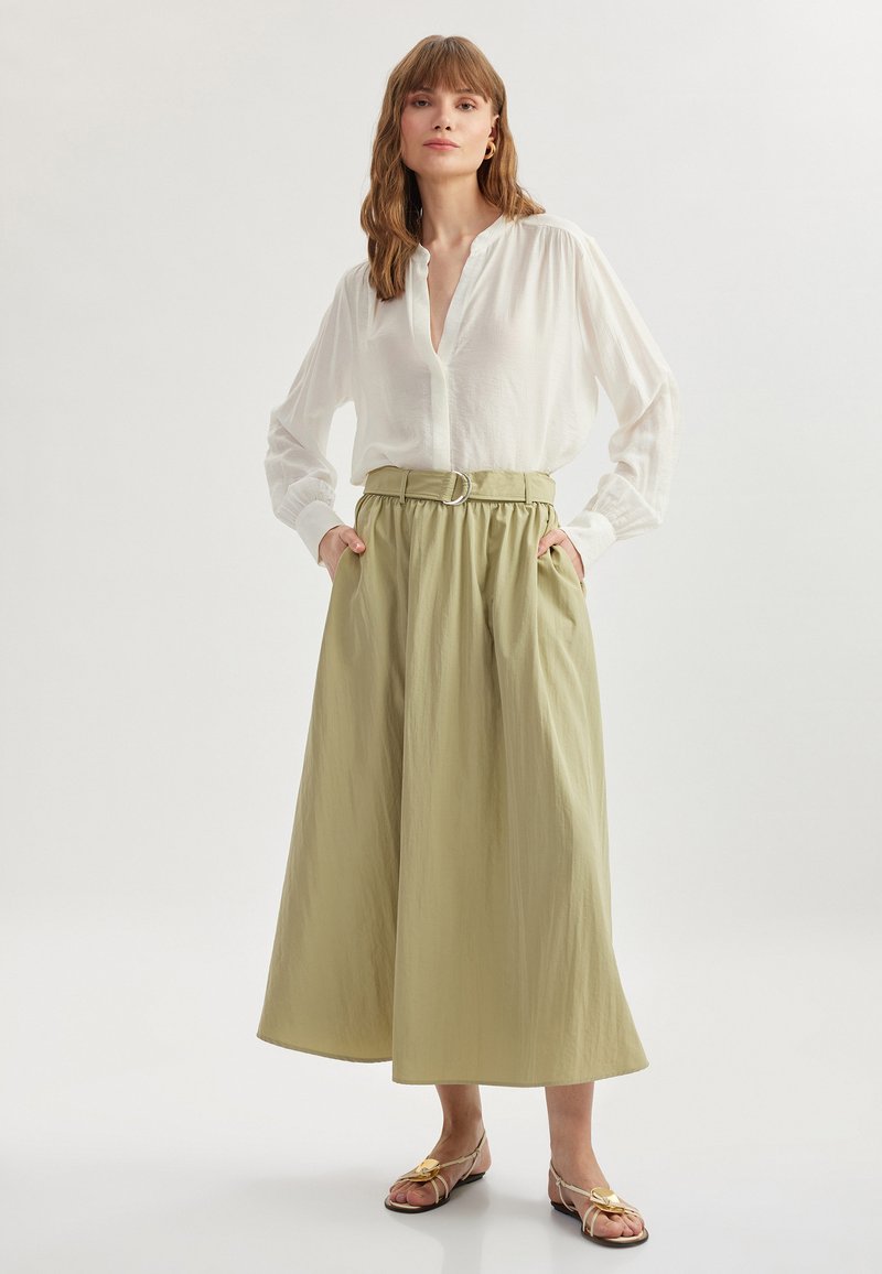 Falda ligera y larga de color verde oliva con cinturilla elástica y detalle de hebilla plateada, combinada con una camisa blanca de botones y sandalias de tiras.