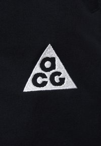 Črn material z belim, vezenim trikotnim logotipom, na katerem sta črki "a" in "cc" v opazni, zaobljeni pisavi. Gladka tekstura z jasnim šivanjem.