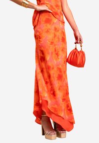 Jupe maxi florale orange vif avec un design fluide et un ourlet asymétrique, associée à un sac à main orange assorti et des talons compensés.