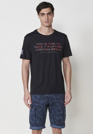 Schwarzes Baumwoll-T-Shirt mit roten und weißen Schriftzügen, kombiniert mit navyblauen Cargo-Shorts mit einem subtilen Muster und hochgerollten Säumen.