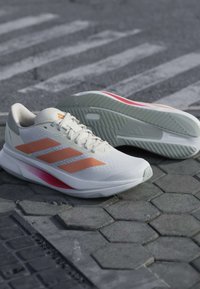 Chaussures de course Adidas blanches et orange posées sur des dalles hexagonales grises près d'un passage piéton.